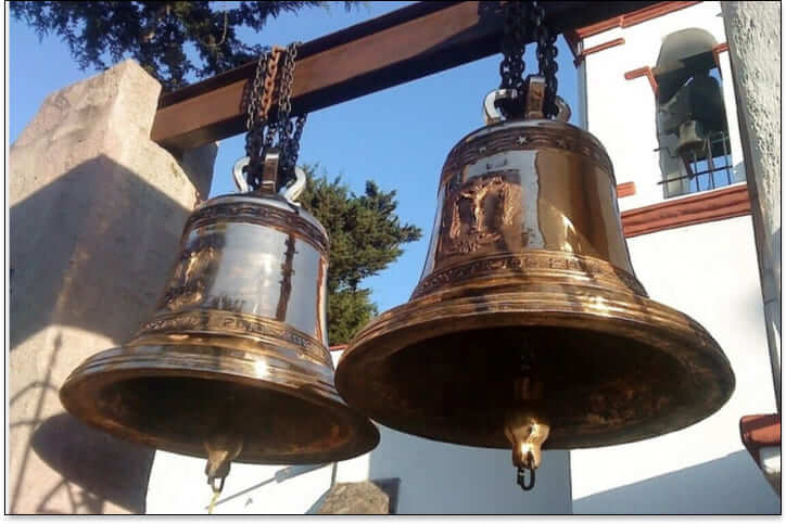 Dos campanas de bronce instaladas en una estructura metálica frente a una iglesia.