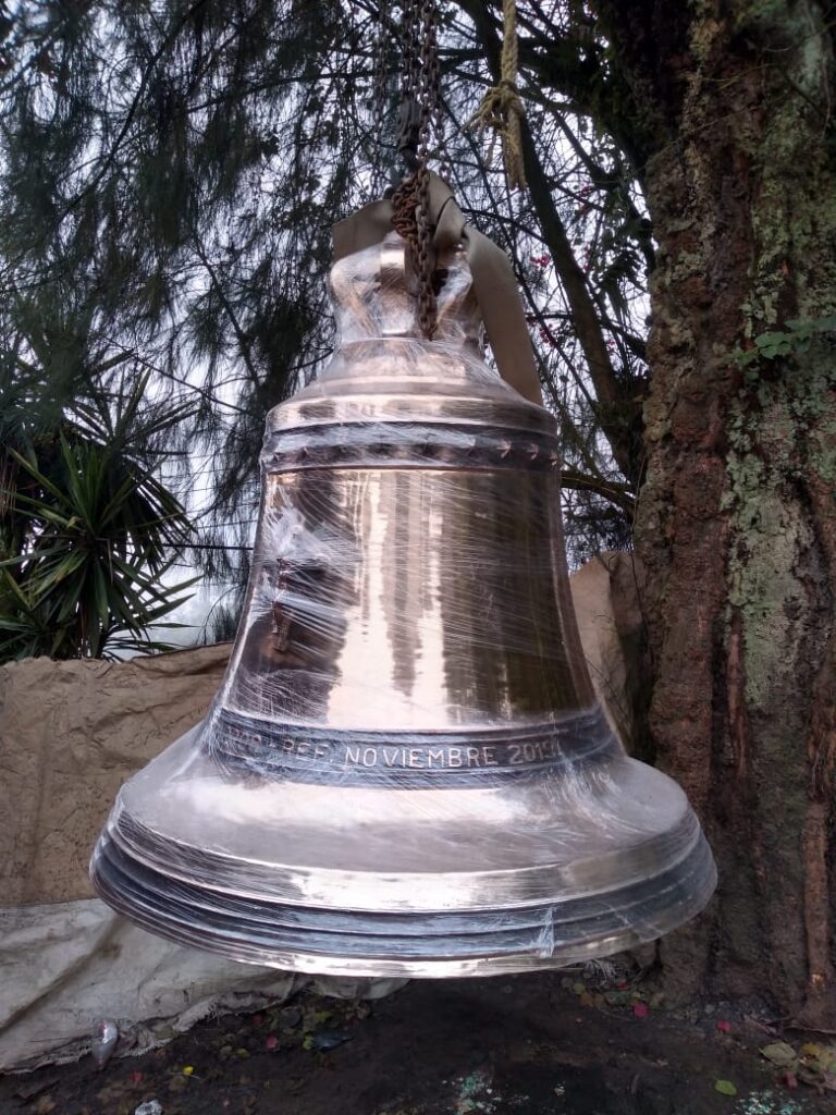 Campana de bronce envuelta en plástico, colgada de una cadena junto a un árbol.